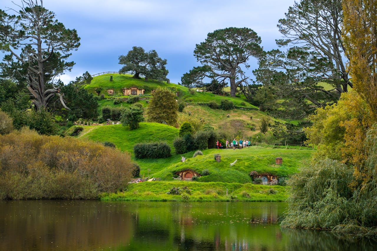 about-us-img Lush green landscape of Hobbiton with iconic hobbit holes and serene lake reflecting the vibrant scenery.
